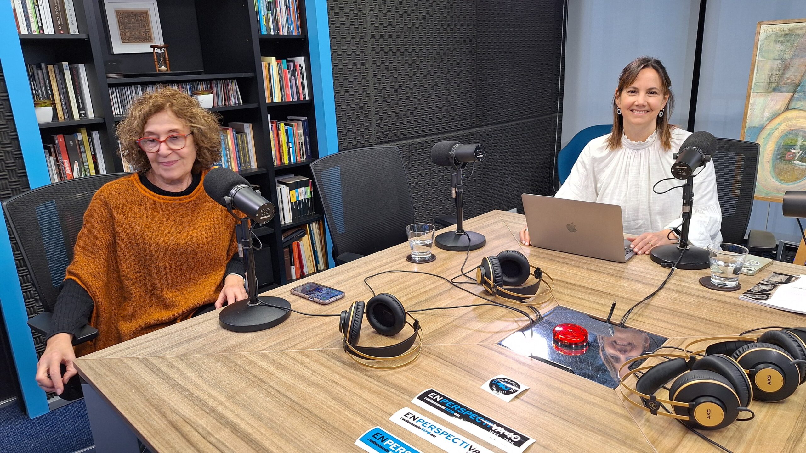 dos mujeres en estudio de radio sentadas en una mesa rectangurar frente a sus microfonos.